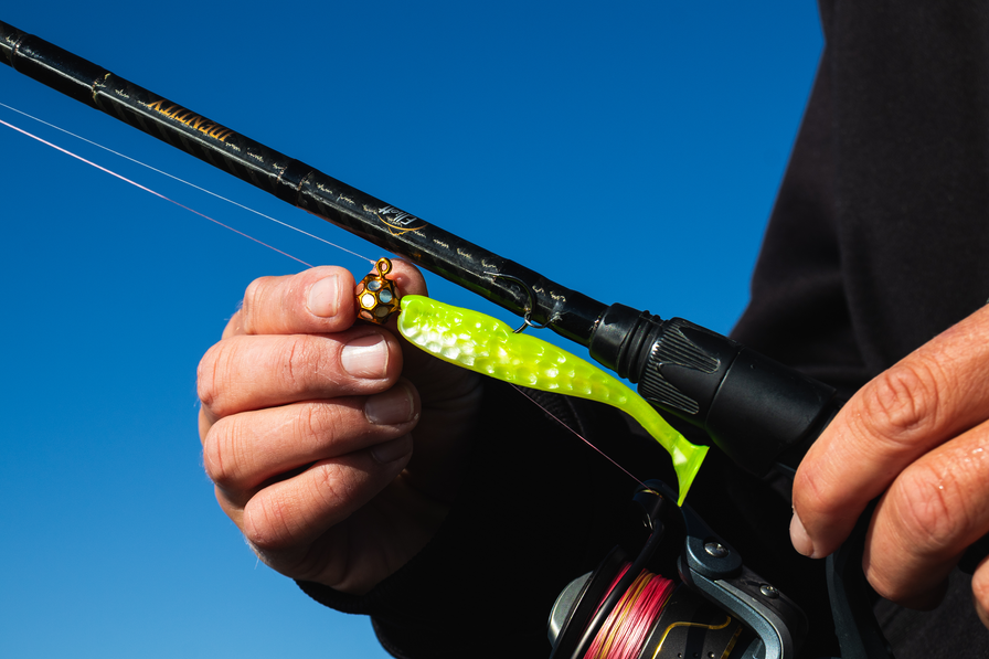 elliott identity fishing rod with a close-up of a hand holding a gold sho-w jig with chartreuse hammered plastic against a clear blue sky.